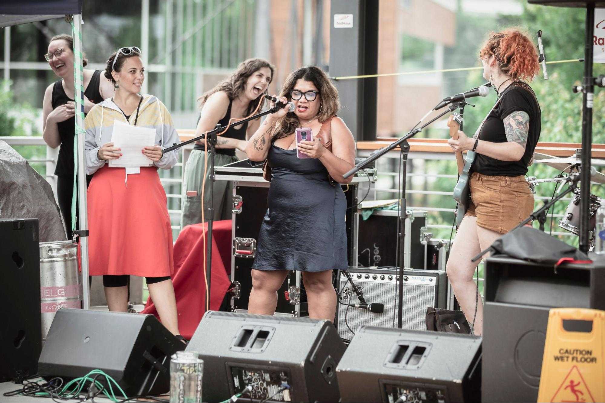 Five women standing on a stage surrounded by audio equipment